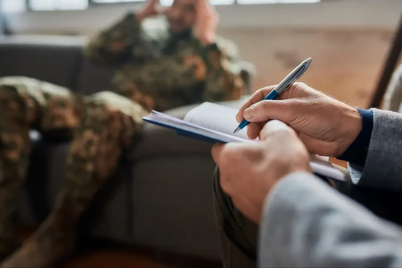 Service member in camouflage seated on a couch while a clinician takes notes