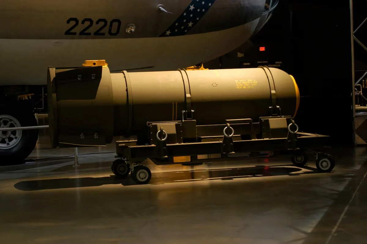 Cylindrical nuclear bomb on a wheeled trolley beneath a bomber marked 2220 in a museum hangar