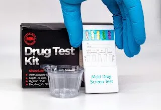 Gloved hand holding a multi-drug screen test beside a drug test kit box and specimen cup