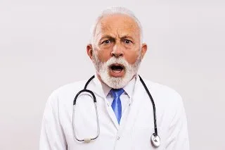 Older doctor in a white coat and stethoscope reacting with an alarmed, open-mouthed expression