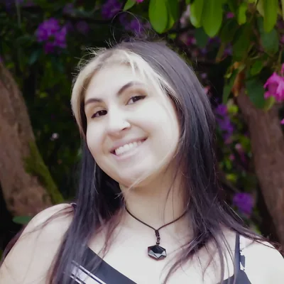 Kayleigh F, a smiling young woman with long dark hair, standing near flowering shrubs.