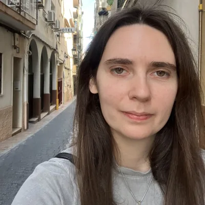 Kelly M, a woman with long brown hair, photographed on a narrow European street.